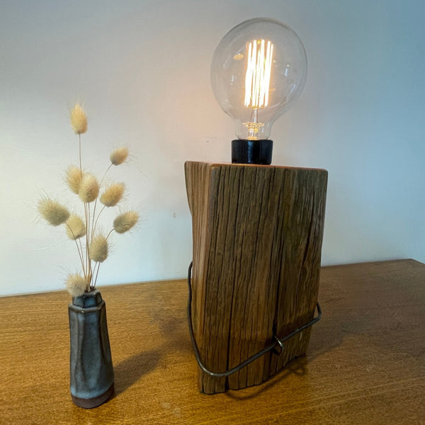 Wood table lamp handcrafted by shades at grays from vintage tōtara fence post,view of front and partial side with replica edison bulb, lit.