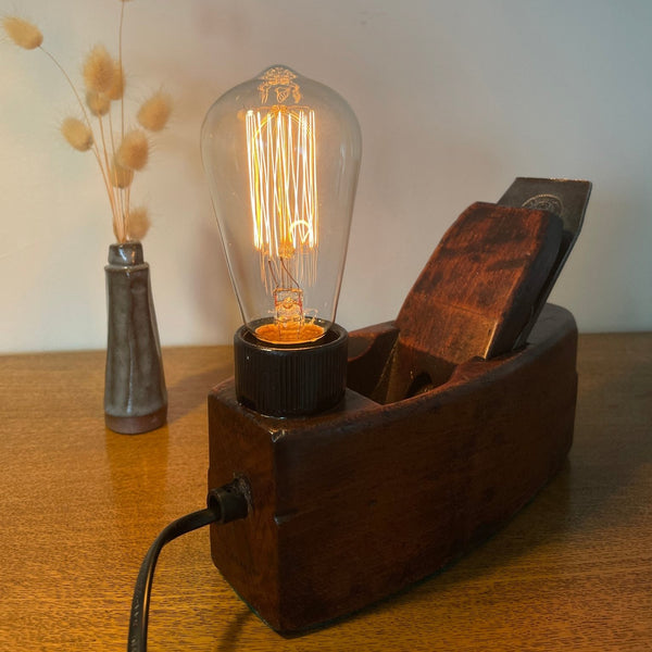 Wood table lamp crafted from authentic carpenters plane, dating back 100 plus years, with replica edison bulb, lit, back view with black lead, sold by shades at grays.