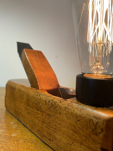 Wood table lamp crafted from authentic carpenters plane, with replica teardrop bulb, lit, side view with sold by shades at grays