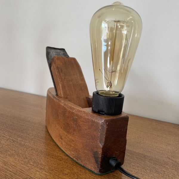 Upcycled carpenter's plane into wood table lamp by shades at grays with replica edison bulb, lit, back view with black lead.