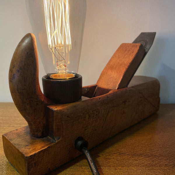 Wood table lamp crafted from authentic carpenters plane, with replica teardrop bulb, lit, close up of back view with black lead by shades at grays