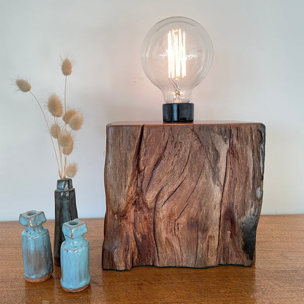 Vintage wood table lamp crafted from upcyled wharf timber with addition of edisn bulb, lit and authentic black and eroded markings, by shades at grays, front view.