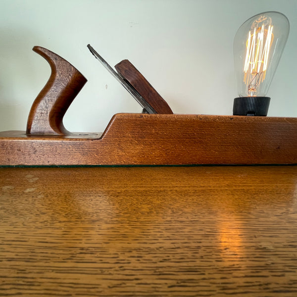 Vintage carpentry wood plane table lamp from Shades at Grays, partial front view with edison bulb, lit.