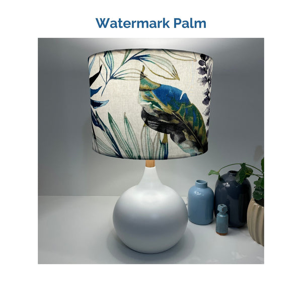 Watermark palm fabric on white touch lamp, lit, with large blue green feather at front, crafted by shades at grays.