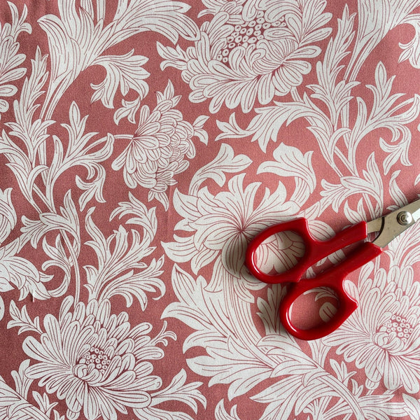 William Morris wandle chrysanthemum fabric sample with white blooms and leaves on a rose pink background, red scissors on fabric for colour comparison.
