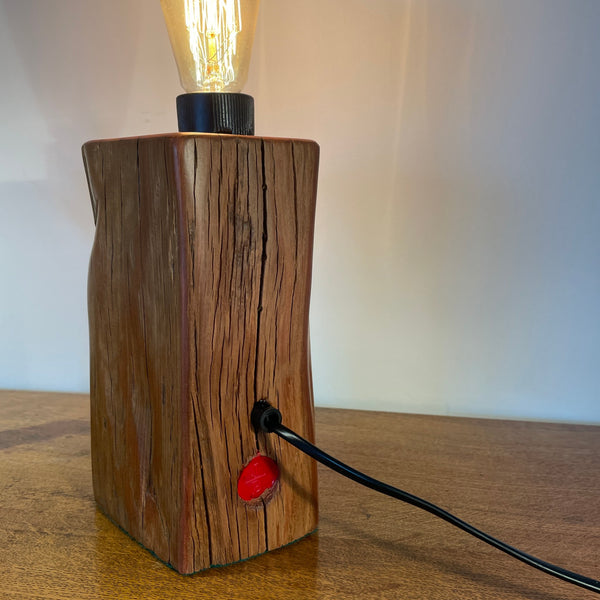 Wood table lamp handcrafted by shades at grays from tōtara fence post, with original staple and edison bulb, lit, back view.