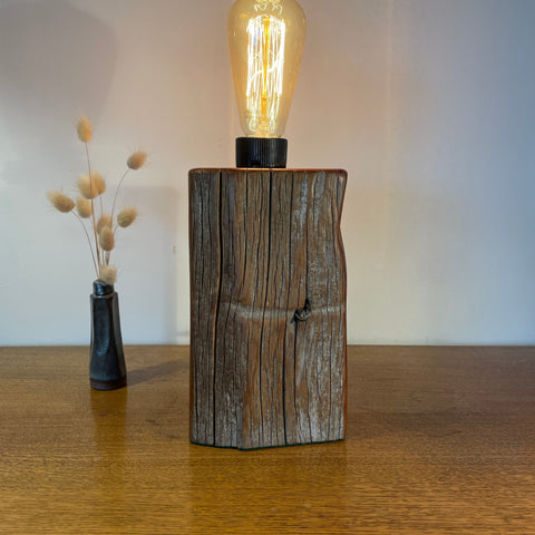 Wooden lamp with a lit bulb on a wooden surface, next to a vase with dried plants.