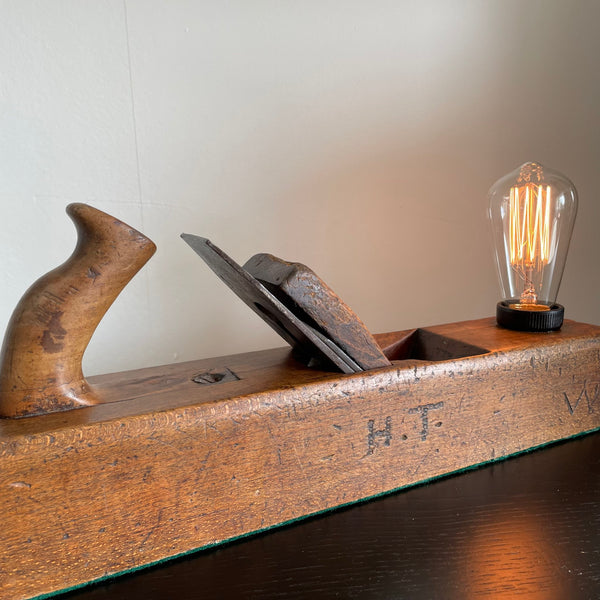 Wooden table lamp crafted from vintage carpenter's plane with owner's initials carved in to cleaned and polished side, by shades at grays.