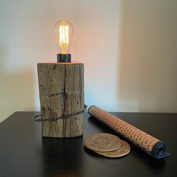 Wood table lamp crafted from reclaimed tōtara post with authentic barbed wire and the adition of an edision bulb, lit, by shades at grays.