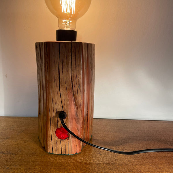 Wood table lamp handcrafted by Shades at Grays from authentic tōtara post, edison bulb, lit, back view with black lead and red makers brand.