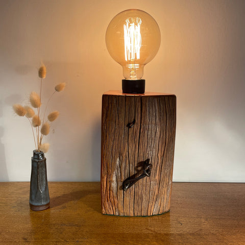 Wooden lamp with a light bulb on a wooden surface, next to a vase with dried plants.
