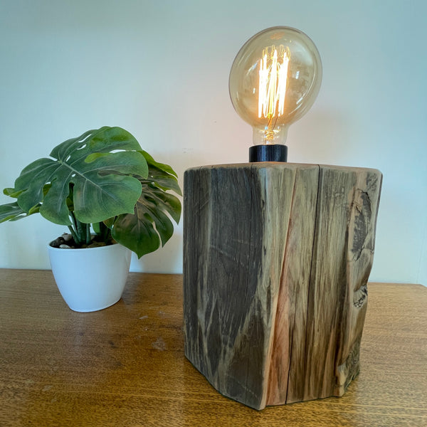 Wood table lamp handcrafted by shades at grays from authentic tōtara post, angled front view of exposed, polished wood grain and lit bulb.