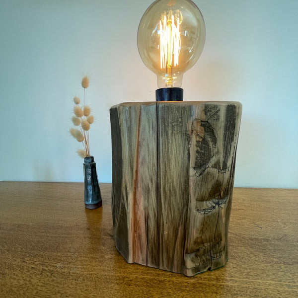 Wood table lamp handcrafted by shades at grays from authentic tōtara post, angled side view of exposed, polished wood grain and lit bulb.