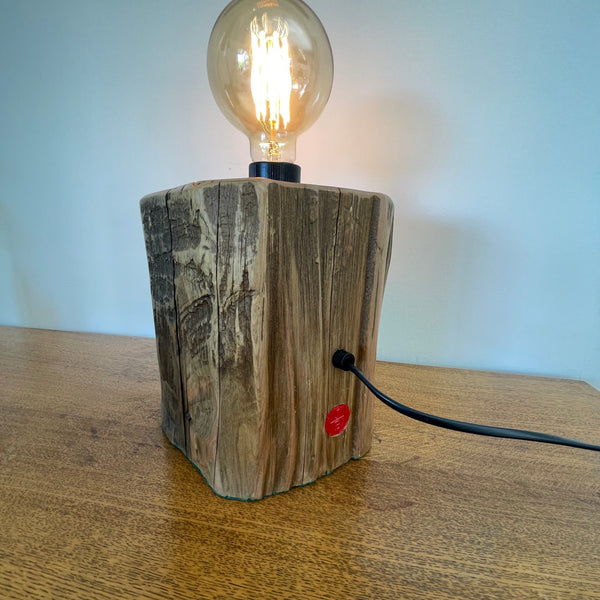 Wood table lamp handcrafted by shades at grays from authentic tōtara post, back view of exposed, polished wood grain and lit bulb.