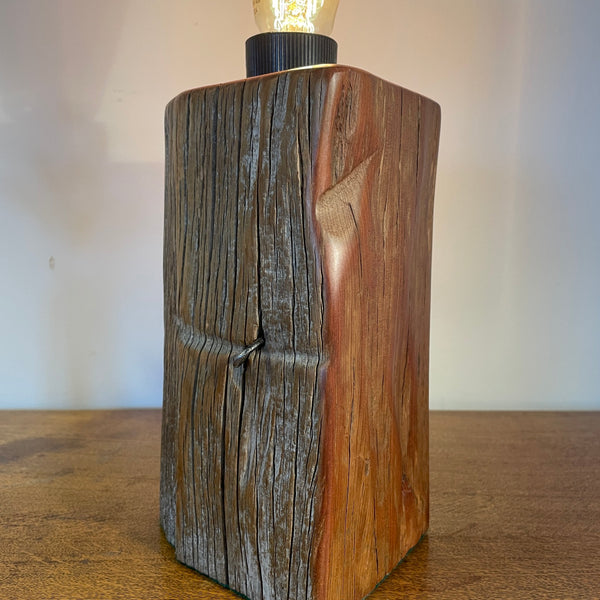 Wood table lamp handcrafted by shades at grays from tōtara fence post, with original staple and edison bulb, lit, front and side view.