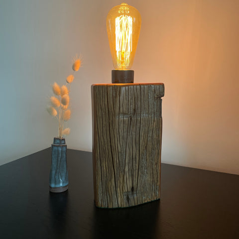 Wooden lamp with a lit bulb on a dark surface next to a vase with dried flowers, handcrafted by shades at grays.