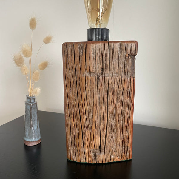 Wooden table lamp with a light bulb on a dark surface next to a vase with dried plants.