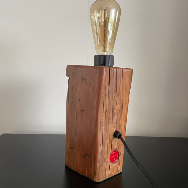 Wooden lamp with a vintage-style light bulb, black lead and makers red brand, on a dark surface.