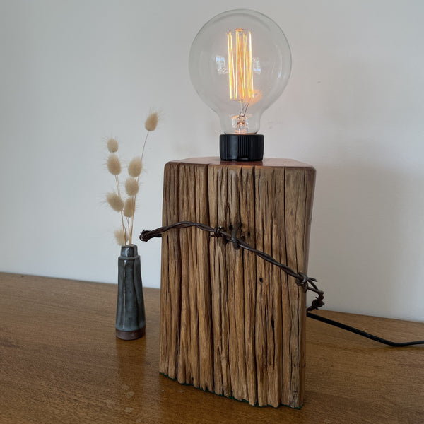 Wood table lamp handcrafted from vintage tōtara post with retained original barbed wire and replica edison bulb, lit, fron view. Made by shades at grays.