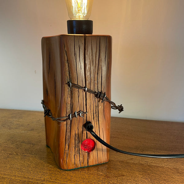 Wood table lamp with polished grain, original wire and replica edison light bulb, lit, back view by shades at grays.
