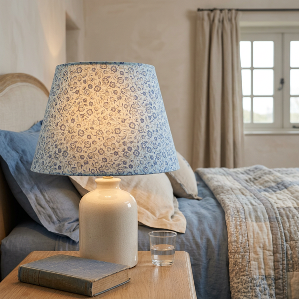 Lit tapered lampshade in French General Renoncule Ciel Blue small flower fabric on a light blue ground, on a cream ceramic base beside a bed.