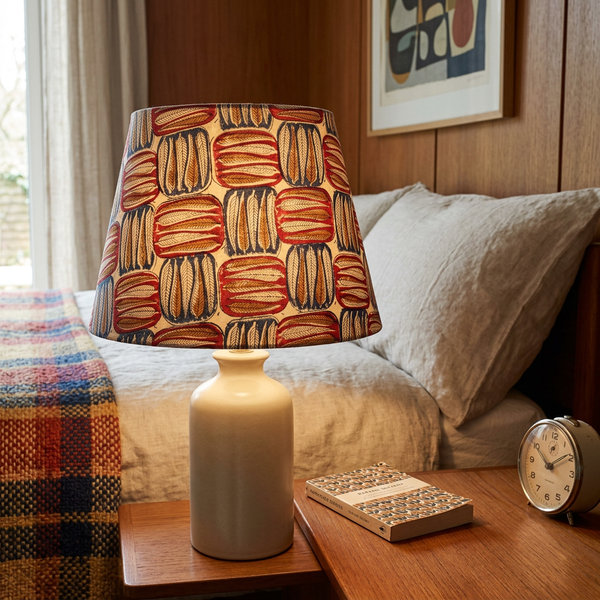 Lit tapered lampshade in Block Leaf Spice fabric on a cream ceramic base beside a bed.