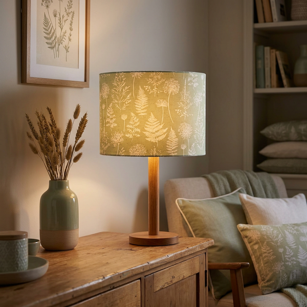 Lit drum lampshade in Meadow Green fabric on a timber table lamp base in a cosy living room.
