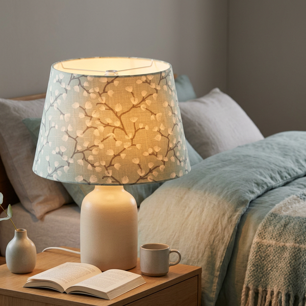 Lit tapered lampshade in Spring Buds Seafoam fabric on a cream ceramic base beside a bed.