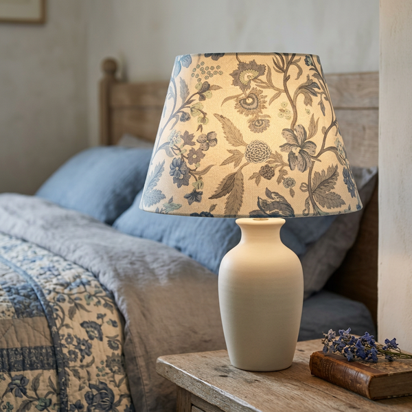 Lit tapered lampshade in French General Parterre Pearl French Blue fabric on a cream ceramic base beside a bed.
