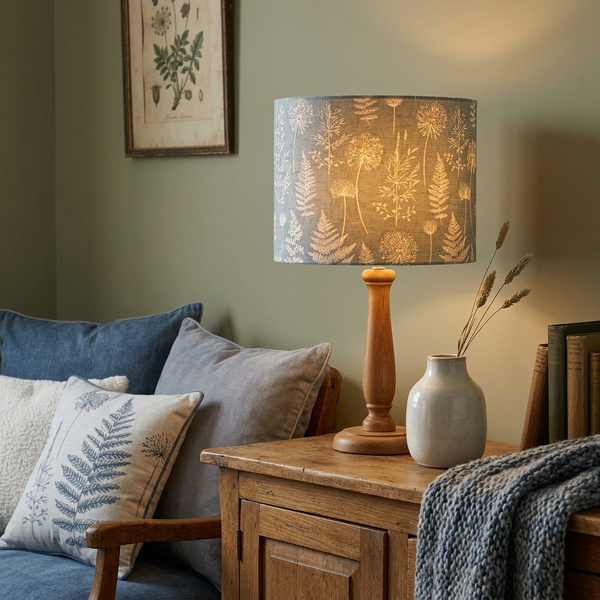 Lit drum lampshade in Meadow Grey Denim fabric on a timber table lamp base in a cosy living room.
