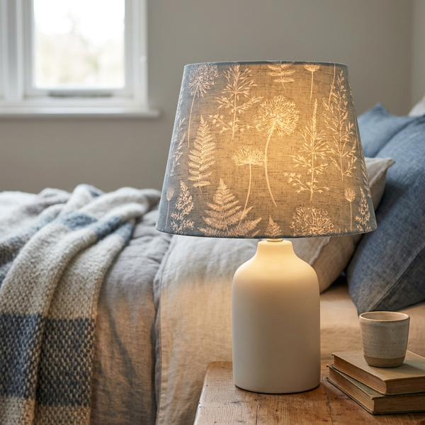 Lit tapered lampshade in Meadow Grey Denim fabric on a cream ceramic base beside a bed.