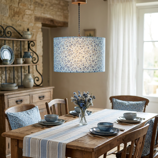 Drum ceiling pendant in French General Renoncule Ciel Blue small flower fabric on a light blue ground, glowing above a wooden dining table.