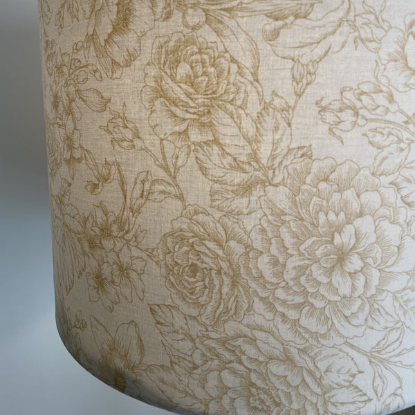 Close up of large drum fabric light shade with flowers etched in gold on cream background, lit.