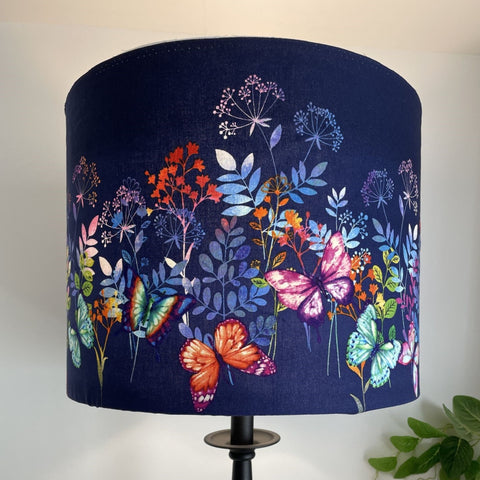 Dark indigo background with multi colour butterflies and foliage on large drum lamp shade, lit.