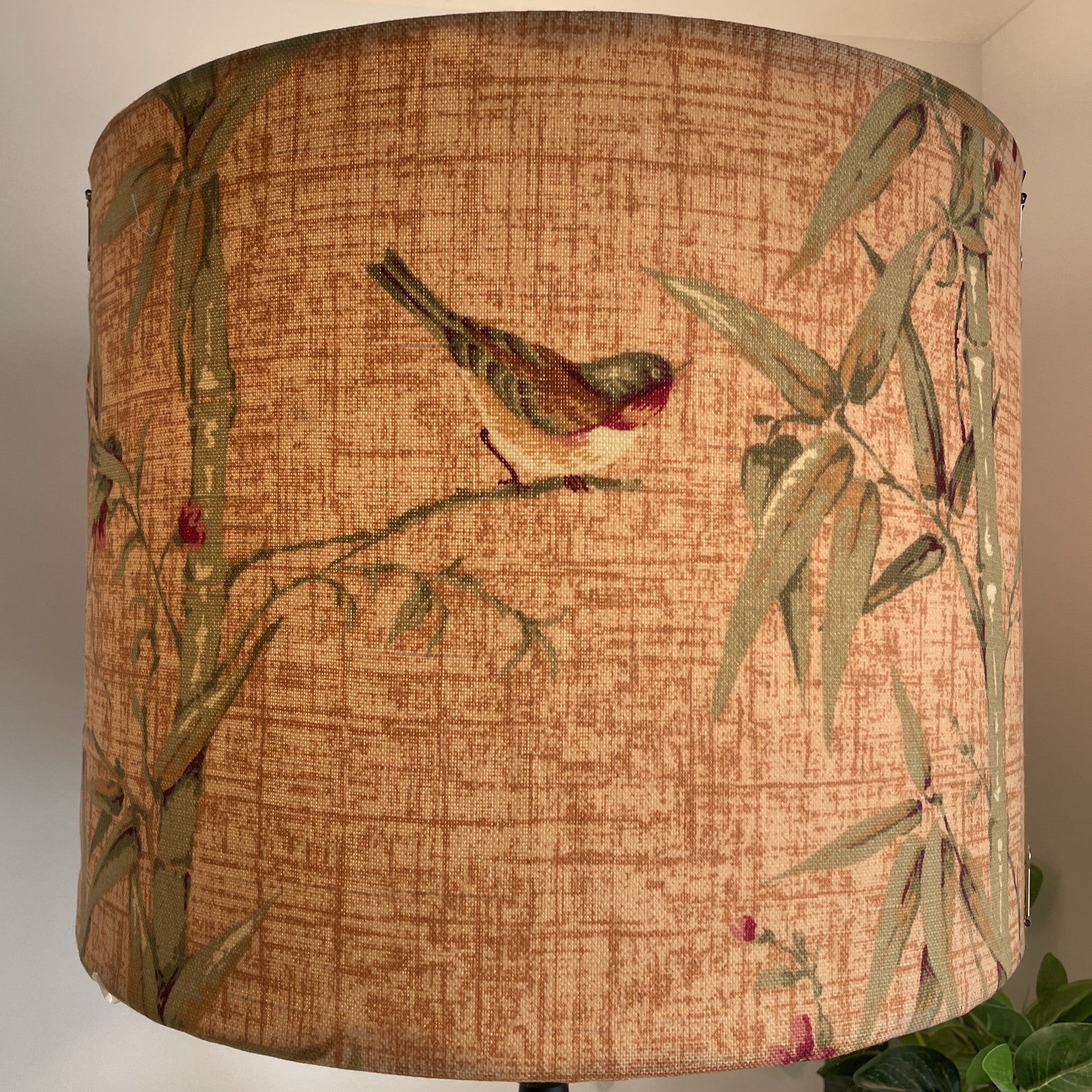 Delicate bird perched on bamboo on large drum lamp shade, lit.