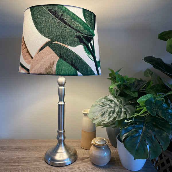 Handcrafted fabric lampshade with large bold green and mushroom coloured leaves on crisp white background, lit, on brushed chrome lamp stand.