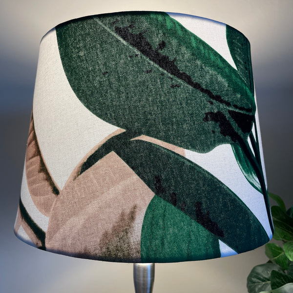 Handcrafted fabric lampshade with large bold green and mushroom coloured leaves on crisp white background, lit, close up.