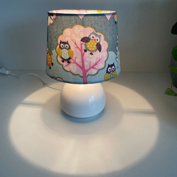Handcrafted small tapered childrens lampshade with owl pattern on white base, lit.