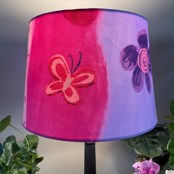 Medium tapered handcrafted fabric lampshade in a variety of pinks with flowers and butterflies. Lit, close up.