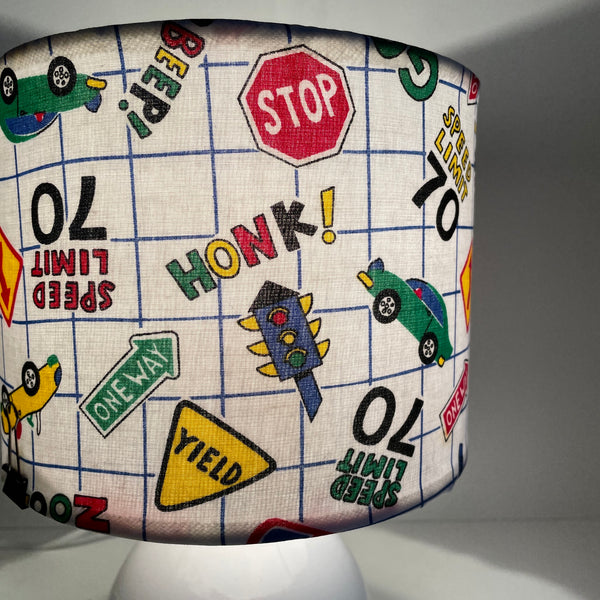 Close up of Small fabric hand crafted lamp shade with fun road signs and cars on white background, lit.