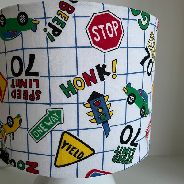 Close up of Small fabric hand crafted lamp shade with fun road signs and cars on white background, unlit, made by Shades at Grays.