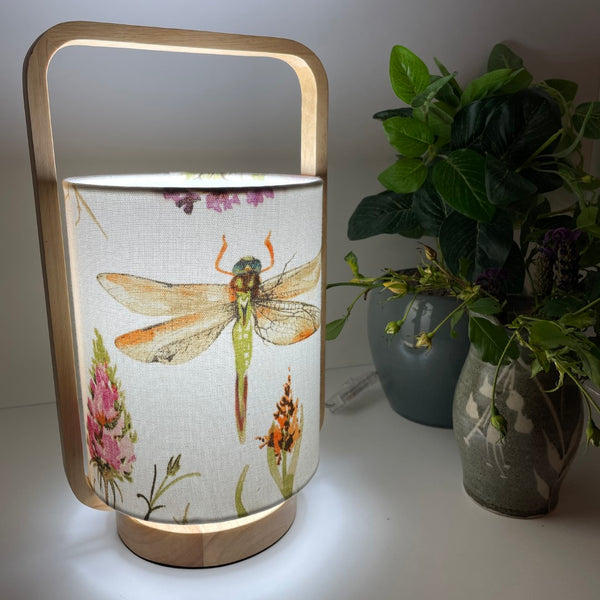 Natural timber table lamp with dragonfly fabric lampshade, lit, angled view.