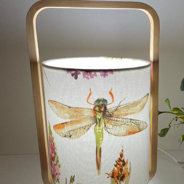 Natural timber table lamp with dragonfly fabric lampshade, lit, close up.