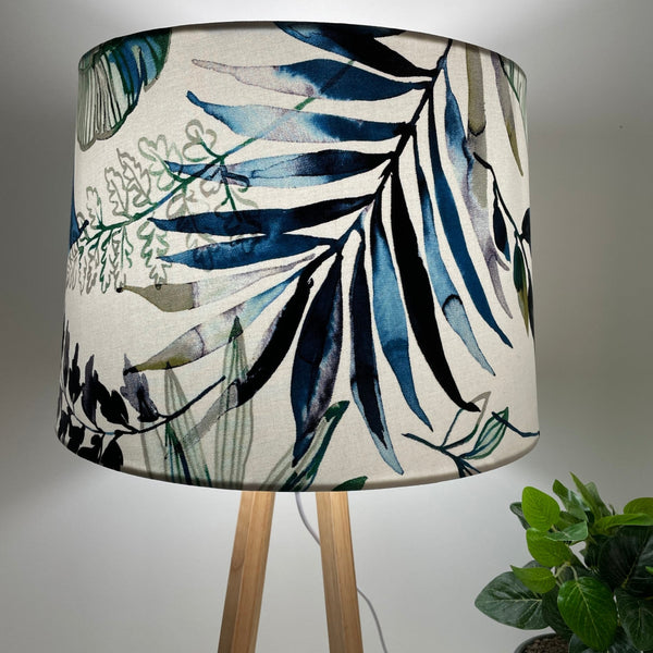Natural wood table lamp with handcrafted fabric lampshade, lit, close up of blue and green foliage on clean white background.