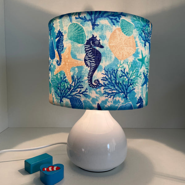 Small handcrafted drum lamp shade with sea horse and star fish fabric on white base, lit.