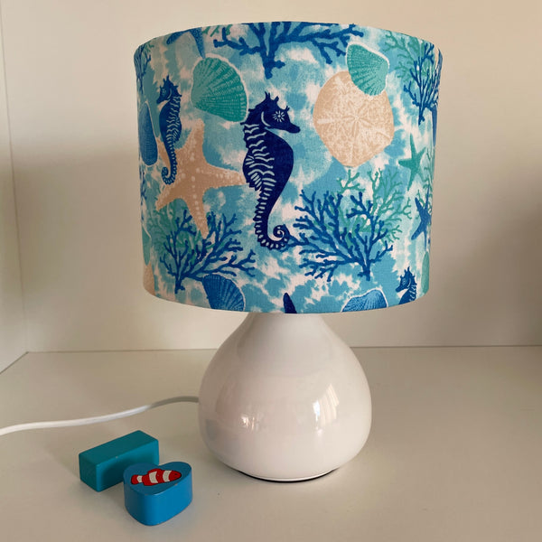 Small handcrafted drum lamp shade with sea horse and star fish fabric on white base, unlit.