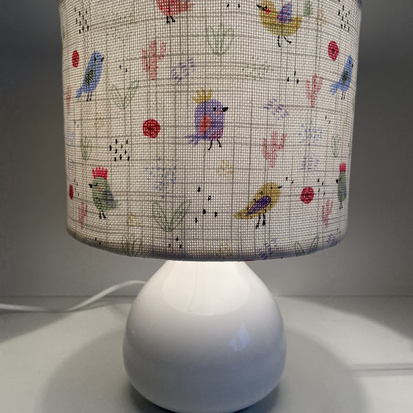 Small hand crafted lamp shade with sweet birdies on a white background lamp shade, lit on small white lamp base.