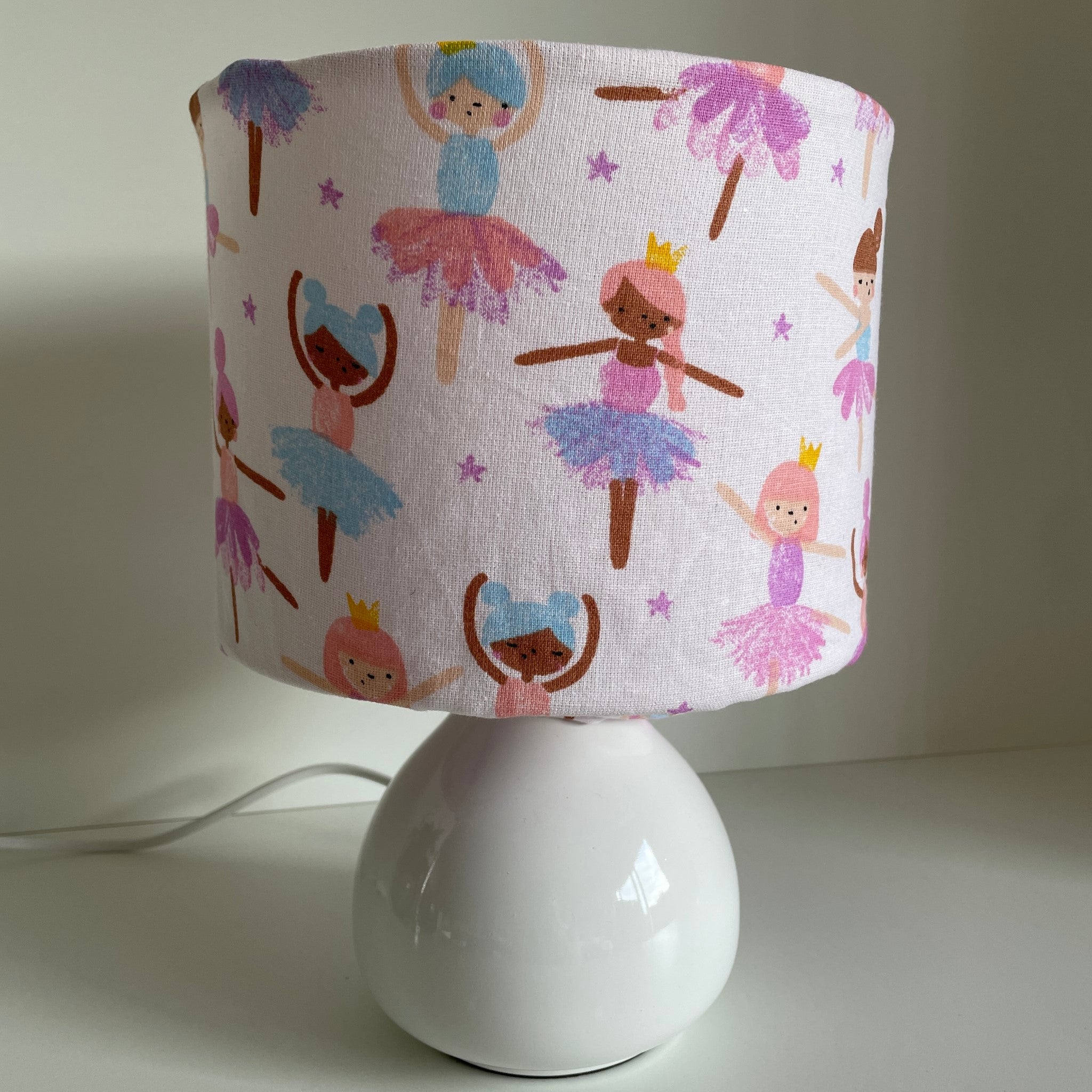 Young ballerina figures dance on a soft cream background, small fabric light shade on small white lamp base.